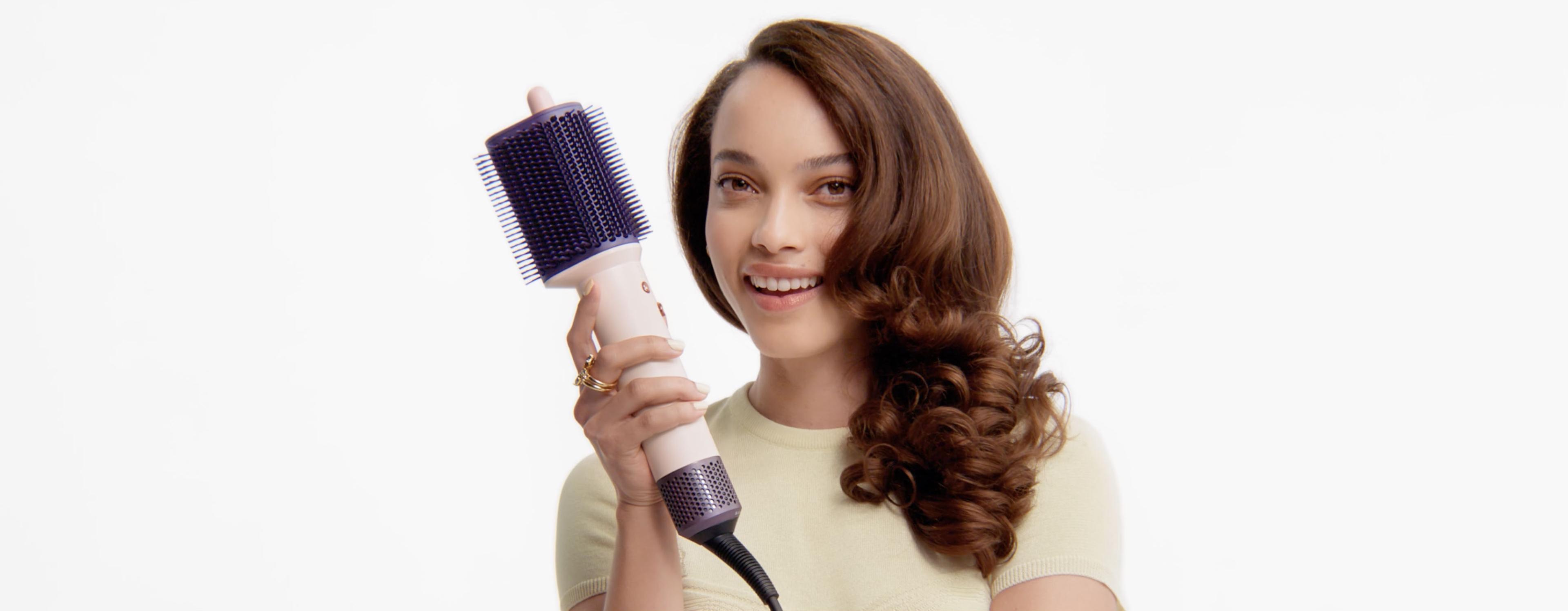 Модель с уложенными волосами держит щётку для укладки Dyson Airsmooth.